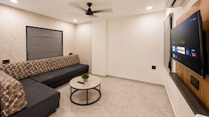 A modern living room with a sectional sofa, a round coffee table, a ceiling fan, and a flat-screen TV mounted on the wall. - Hotel Shree Dwarka Devbhumi Dwarka (Dwarka)