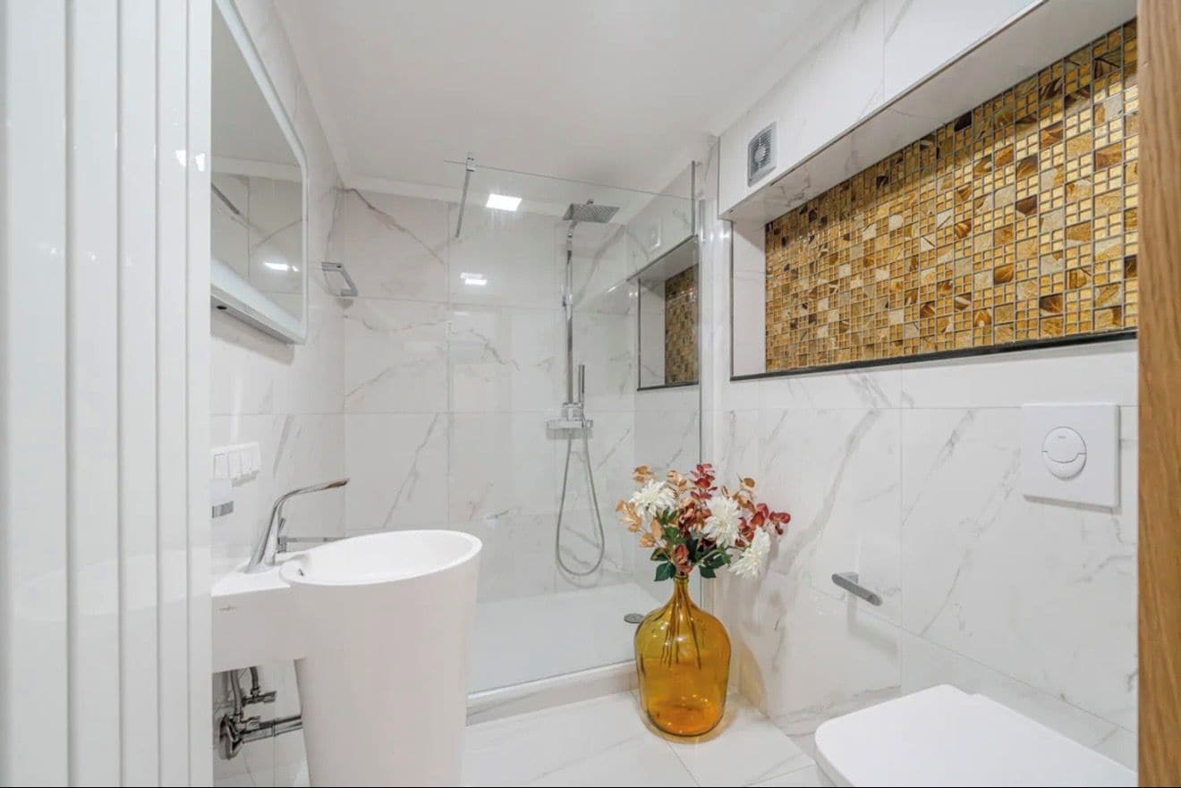 Un baño moderno con un lavamanos blanco, ducha con cerramiento de vidrio y pared de mosaico.