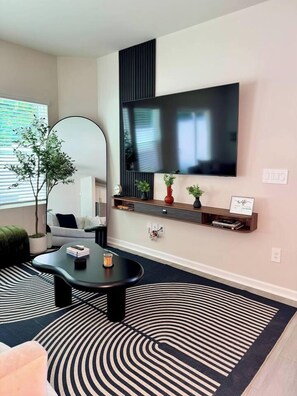 A modern living room with a flat-screen TV mounted on the wall, a black coffee table, a sofa, a large mirror, and a patterned rug.