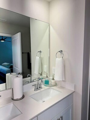 A modern bathroom with a large mirror, two sinks, and a visible door leading to a bedroom.