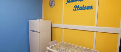 A compact kitchen with a white refrigerator, a small table with chairs, and a yellow partition with 'Maison Platone' written on it.