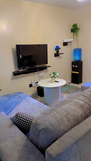 A living room with a large flat-screen TV mounted on the wall, a round white coffee table with a vase of flowers, a grey sofa with patterned cushions, and a water dispenser on a black cabinet. - Thika road 1 bedroom executive  Airbnb unit located off Thika road at exit 12 (Ruiru)