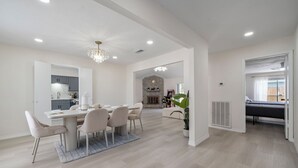Une salle à manger spacieuse avec un haut plafond et un lustre moderne, qui donne sur une cuisine et un salon agrémenté d’une cheminée.