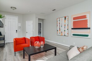 Ein Wohnzimmer mit zwei roten Sesseln, einem schwarzen Couchtisch und einem grauen Sofa. An den Wänden befinden sich zwei abstrakte Gemälde.