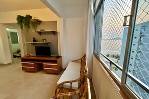 A living room with a TV, wooden furniture, a window with a view of buildings, and a plant on the wall.