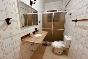 Baño con pared de azulejos blancos, encimera beige, lavabo blanco, inodoro y cabina de ducha.