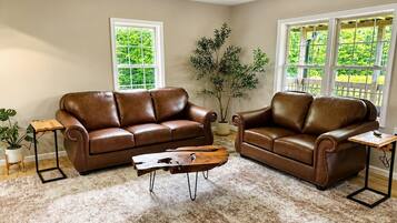 A living room with two brown leather sofas, a wooden coffee table, and a large window providing a view of greenery.