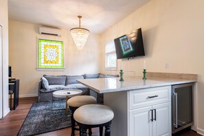 Una cocina moderna con horno empotrado, zona de comedor con sofá y taburetes, una araña y un televisor de pantalla plana montado en la pared.