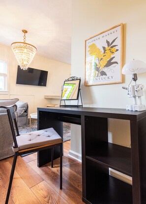 A modern living room with a dark wooden desk, a comfortable chair, a television mounted on the wall, a chandelier, and a framed picture of flowers. - Second-floor 2BR above Clifton's restaurant row | Stained glass, high ceilings | Rain shower, 750 Mbps | Walk everywhere (Louisville)