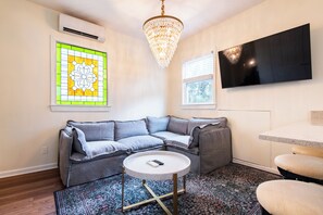 A modern living room with a grey sectional sofa, a round coffee table, a chandelier, a flat-screen TV, and a stained glass window. - Second-floor 2BR above Clifton's restaurant row | Stained glass, high ceilings | Rain shower, 750 Mbps | Walk everywhere (Louisville)