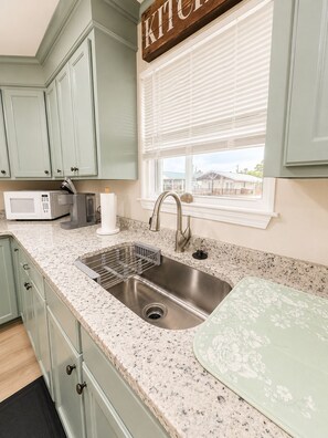 A kitchen with a stainless steel sink, a microwave, and a toaster. There is a window with blinds and a view of houses outside.