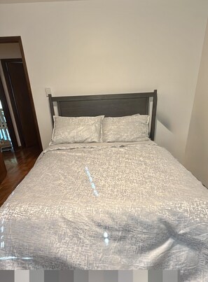 Cama com colcha texturizada e duas almofadas, cabeceira de madeira e parede lisa.