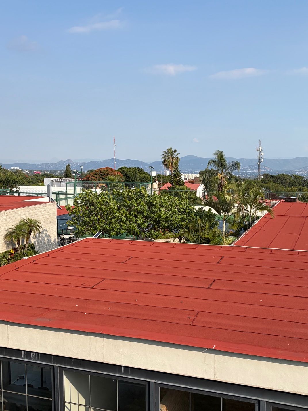 A building with a red roof, surrounded by greenery and palm trees, with a clear sky and distant mountains.