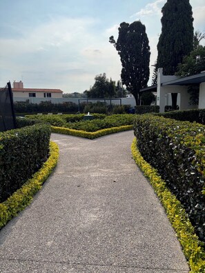 A paved pathway flanked by neatly trimmed hedges and yellow flowers, leading towards a building with a white facade. - Panoramic view of the volcanoes, 1 hour from Taxco and 2.5 hours from the beach (Cuernavaca)