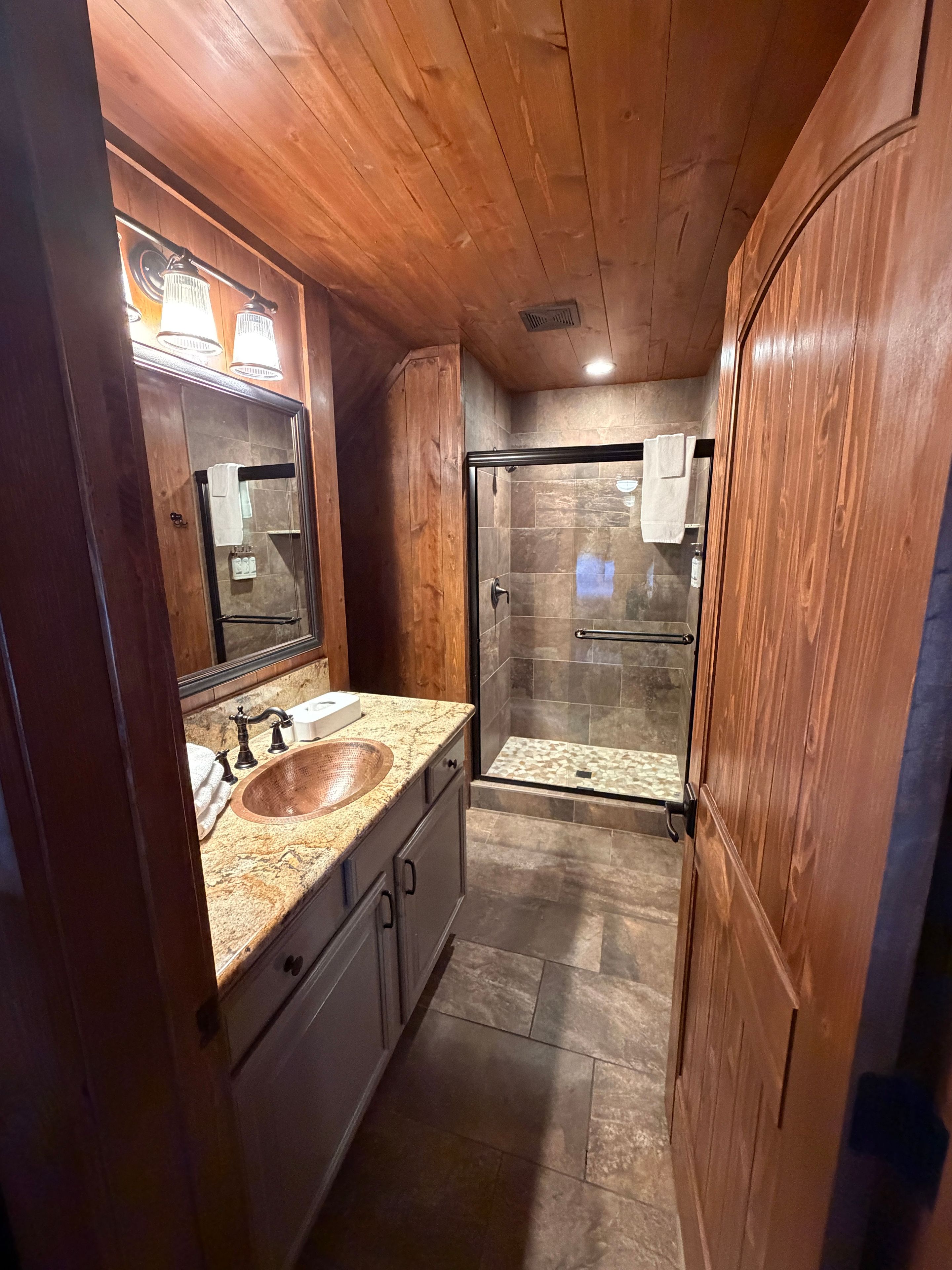 Une salle de bain aux murs lambrissés, dotée d’un lavabo avec un plan de travail en granit, d’une douche en verre et d’un miroir avec des lumières au-dessus.