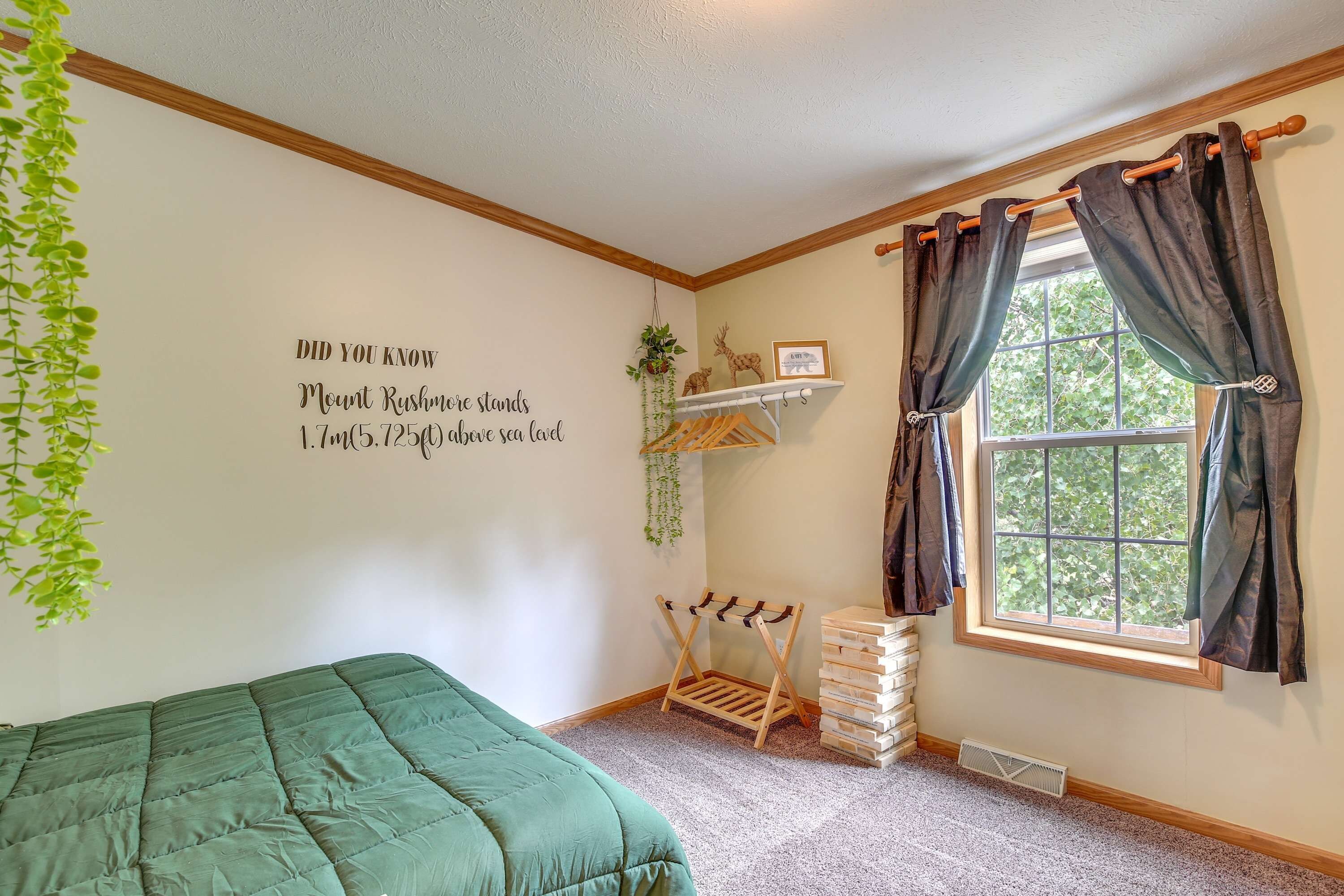 Une chambre avec un lit vert, une étagère fixée au mur ornée de plantes, une fenêtre avec des rideaux et un motif décoratif mural avec du texte.