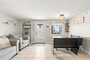 A living room with a sofa, a dining table, and a piano. There is a door with a window, and the room has a tiled floor. - 2 bedroom ocean view condo at Grandview! (Old Orchard Beach)