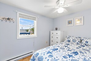 A bedroom with a bed, a dresser, a ceiling fan, a window with a view of a motel, and wall art. - 2 bedroom ocean view condo at Grandview! (Old Orchard Beach)