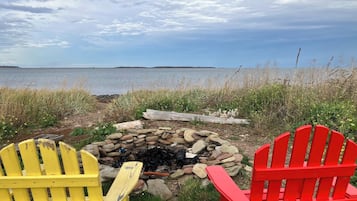 Ein Strandbild mit einem Feuerkorb, umgeben von Steinen, zwei Adirondack-Stühlen und Blick aufs Meer.