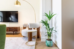 Ein modernes Wohnzimmer mit rundem Tisch, zwei Stühlen, einem Fernseher, einer Stehlampe, einer grünen Pflanze und einem weißen Teppich.