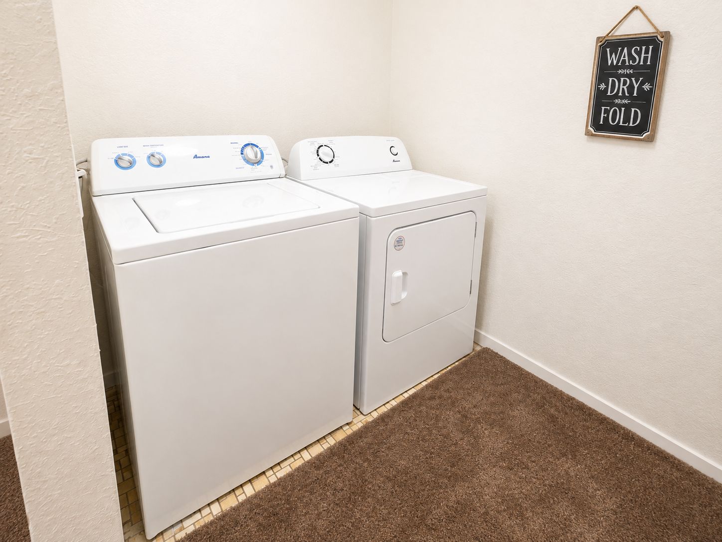 A laundry room with a front-loading washer and dryer, a brown carpeted floor, and a wall-mounted sign that reads 'WASH DRY FOLD'.