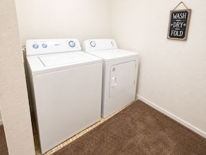 A laundry room with a front-loading washer and dryer, a brown carpeted floor, and a wall-mounted sign that reads 'WASH DRY FOLD'.