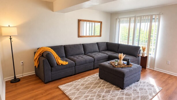 Ein Wohnzimmer mit einem Ecksofa, einem Pouf, einer Stehlampe und einem Fenster mit Jalousien.