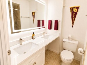 A bathroom with two sinks, a toilet, and a shower. There are towels hanging on a rack and a towel dispenser. - New! CMU Getaway | Near Soaring Eagle & Campus (Mount Pleasant)
