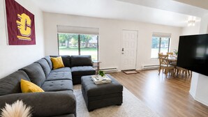 A modern living room with a sectional sofa, ottoman, and a flat-screen TV. - New! CMU Getaway | Near Campus & Soaring Eagle (Mount Pleasant)