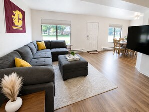 A modern living room with a sectional sofa, ottoman, and a flat-screen TV. - New! CMU Getaway | Near Campus & Soaring Eagle (Mount Pleasant)
