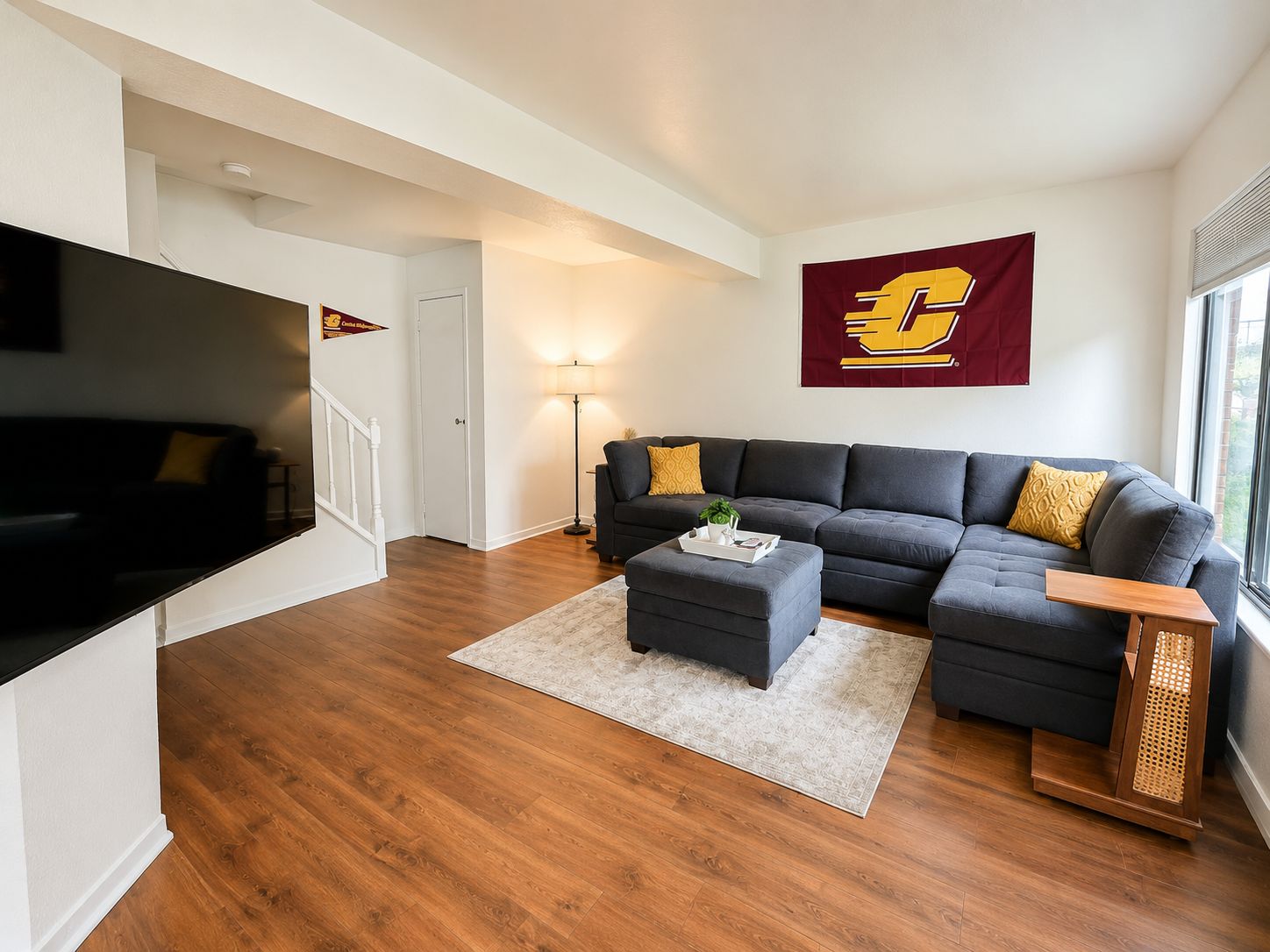 A living room with a dark grey sectional sofa, a small ottoman, a flat-screen TV, a flag, and a staircase.