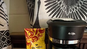 En kaffemaskine med et digitalt ur, der viser 8:38, en pose kaffe mærket 'Gevalia Decof House Blend' og en kop med kaffekapsler.