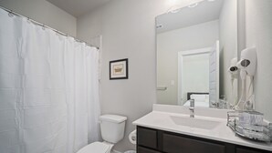 Une salle de bain comprenant des toilettes, un rideau de douche, un tableau encadré au mur, un sèche-cheveux et un lavabo avec un miroir.