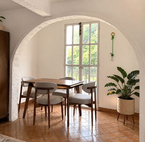 A dining area with a wooden table and chairs, a large window with a view of greenery, and a potted plant on a stand. - Just 5 minutes from the archaeological site! Ideal family apartment with a garden (Malinalco)
