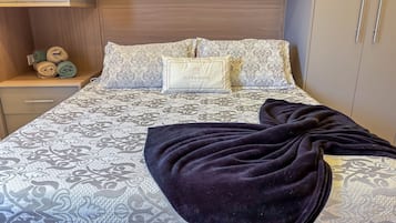Cama bem arrumada com roupa de cama estampada, um travesseiro com a inscrição 'SUPER HOST BRASIL' e um cobertor escuro.