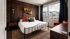 Una camera da letto con un letto, un quadro raffigurante una chitarra, una finestra con tende e vista sugli edifici circostanti.