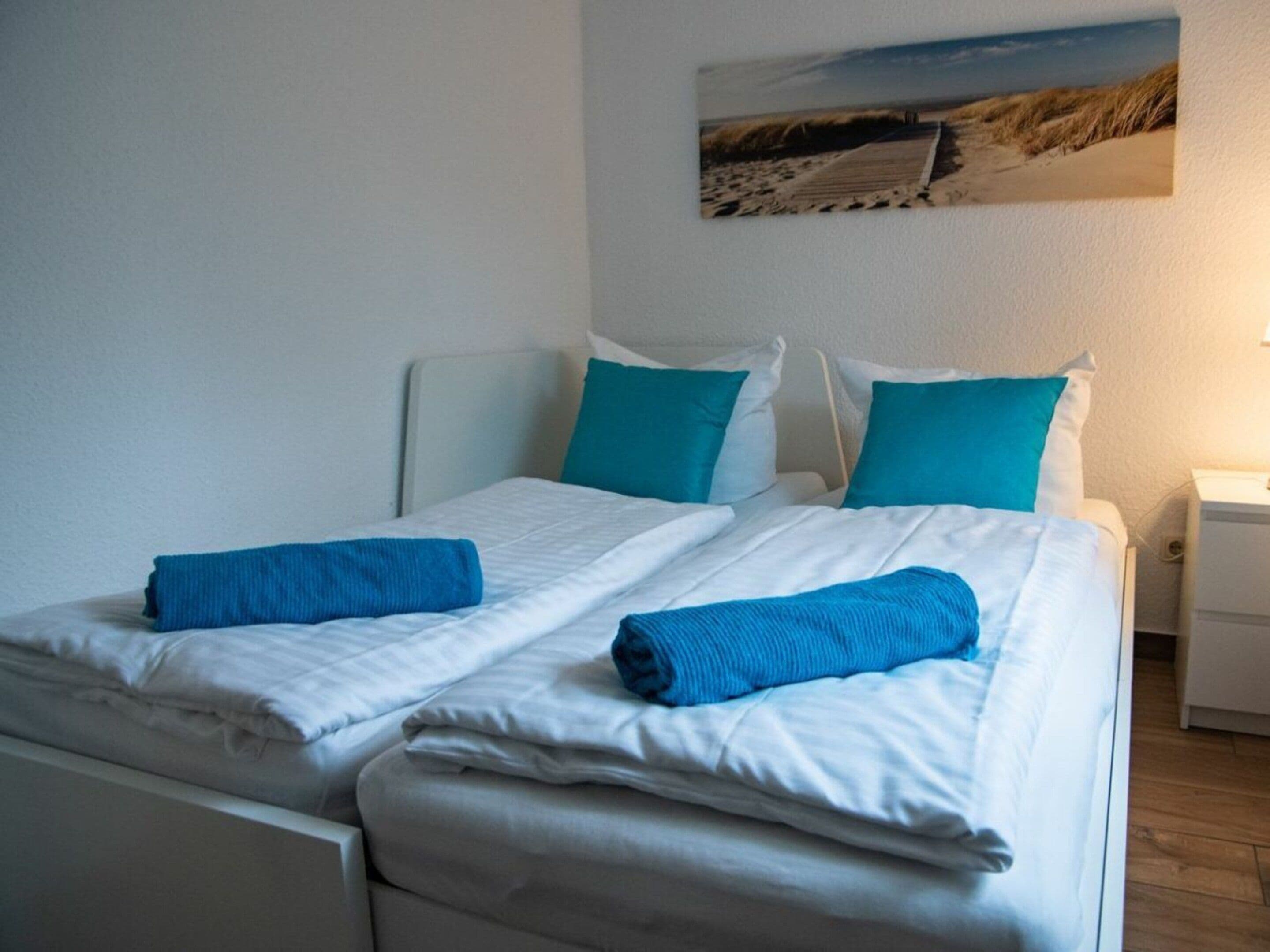 Cama doble con ropa de cama blanca y dos toallas azules, una mesita de noche blanca con lámpara, y un cuadro con motivos playeros.