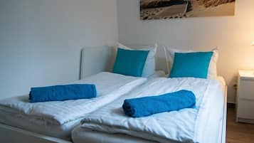Cama doble con ropa de cama blanca y dos toallas azules, una mesita de noche blanca con lámpara, y un cuadro con motivos playeros.