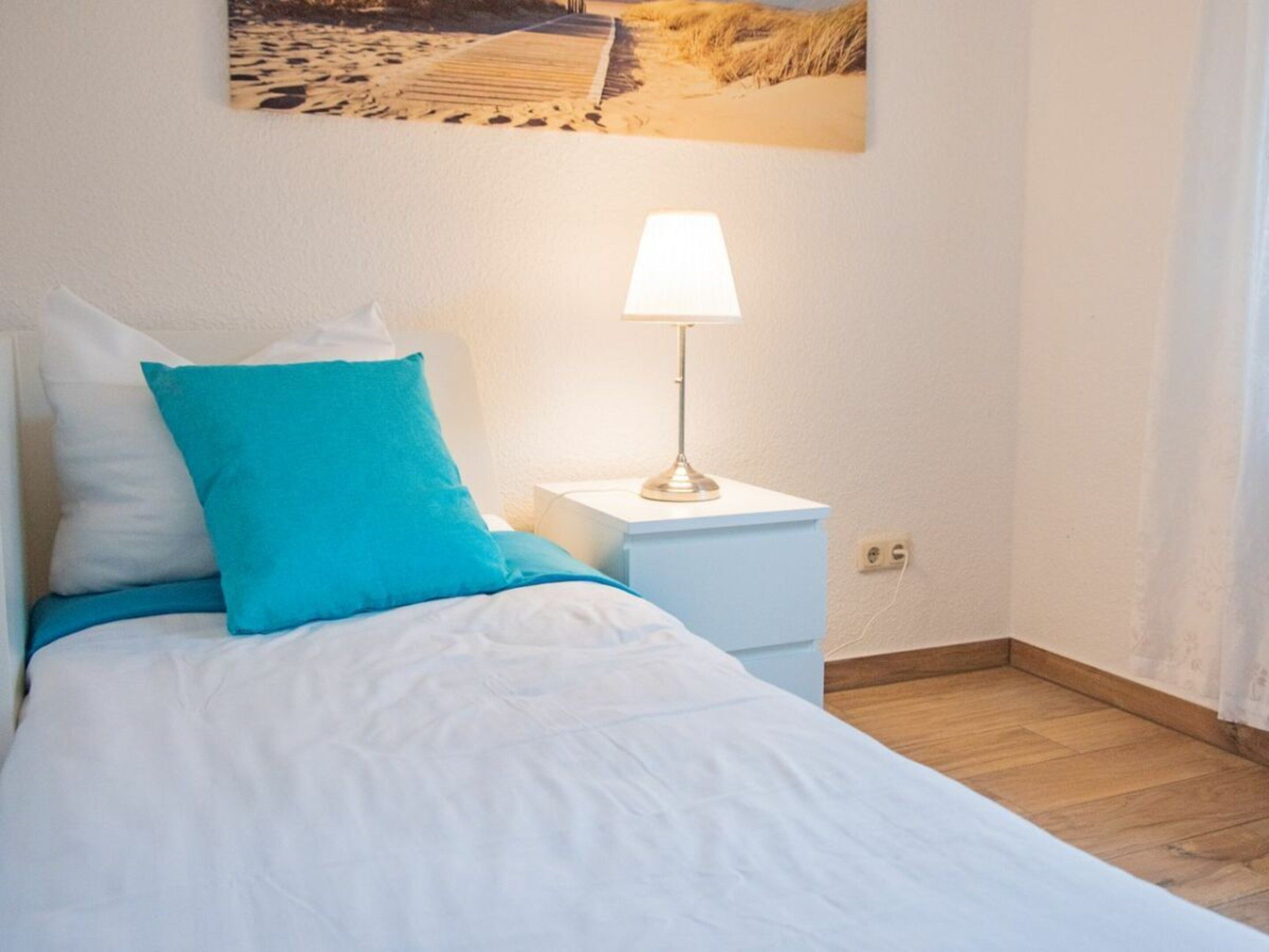 A neatly made bed with white and blue bedding, a bedside lamp, and a picture of a beach scene on the wall.