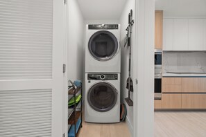 A compact laundry room with two stacked washing machines, a drying rack, and a broom. - Brand-New Furnished Luxury, Sophisticated Turnkey at a Premier Address (North Miami)