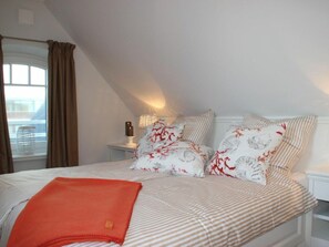 A bedroom with a bed, pillows, a bedside table, a lamp, and a window with curtains. - Holiday Apartment in Sylt With 2 Bedrooms (Sylt)