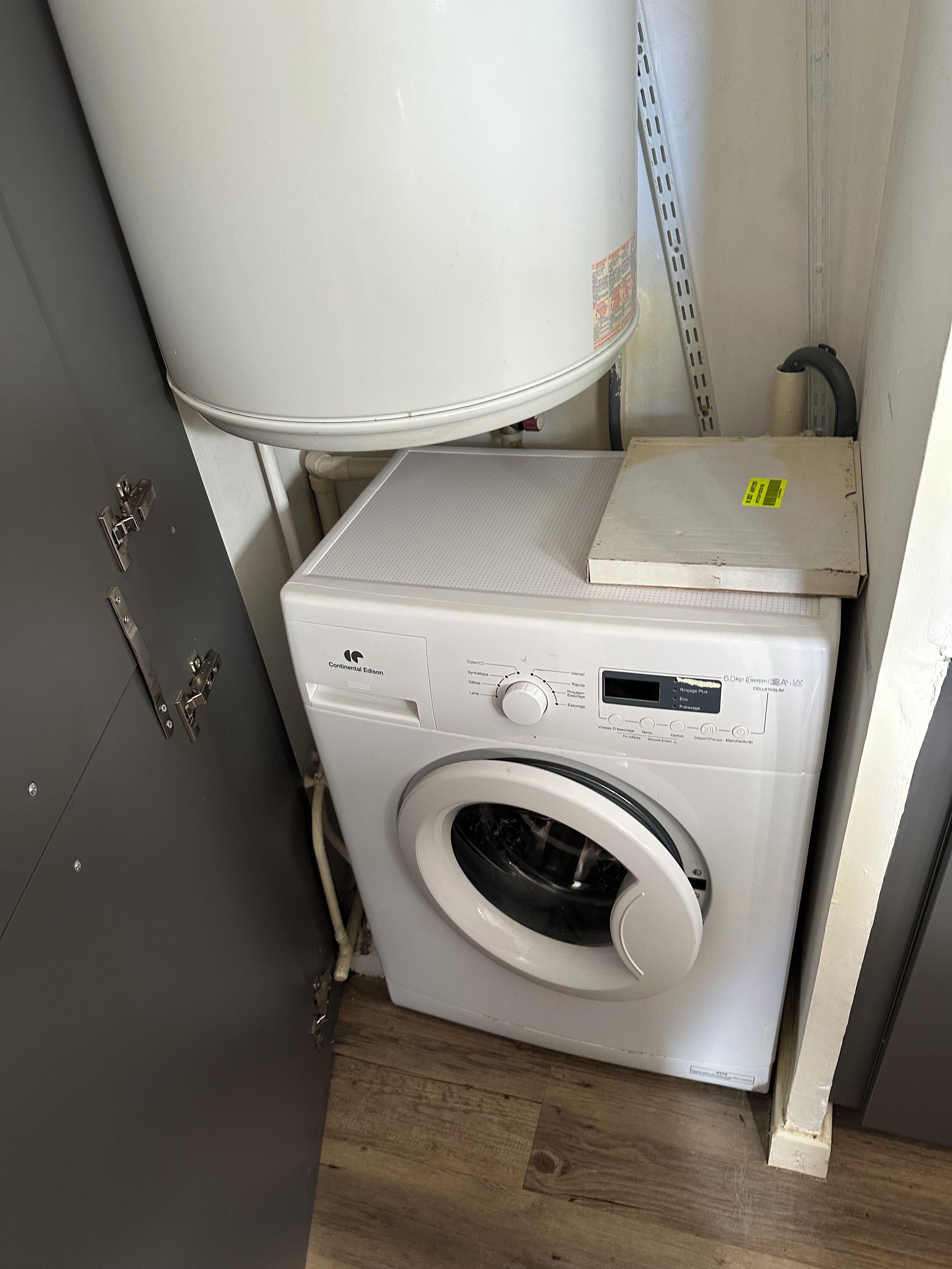Een compacte, witte wasmachine met voorlader, bedieningspaneel en een klein kastje erboven, geplaatst in een smalle ruimte met een grijs kastje en een metalen plank.