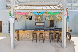 Ein Außenbereich mit Bar, Holztresen und Barhocker. Ein Schild weist darauf hin, dass Glasflaschen und -behälter im Poolbereich nicht erlaubt sind.