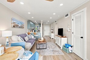 Ein modernes Wohnzimmer mit Sofa, Couchtisch und Fernseher. Es gibt eine Küchenzeile mit Schränken und einem Esstisch.