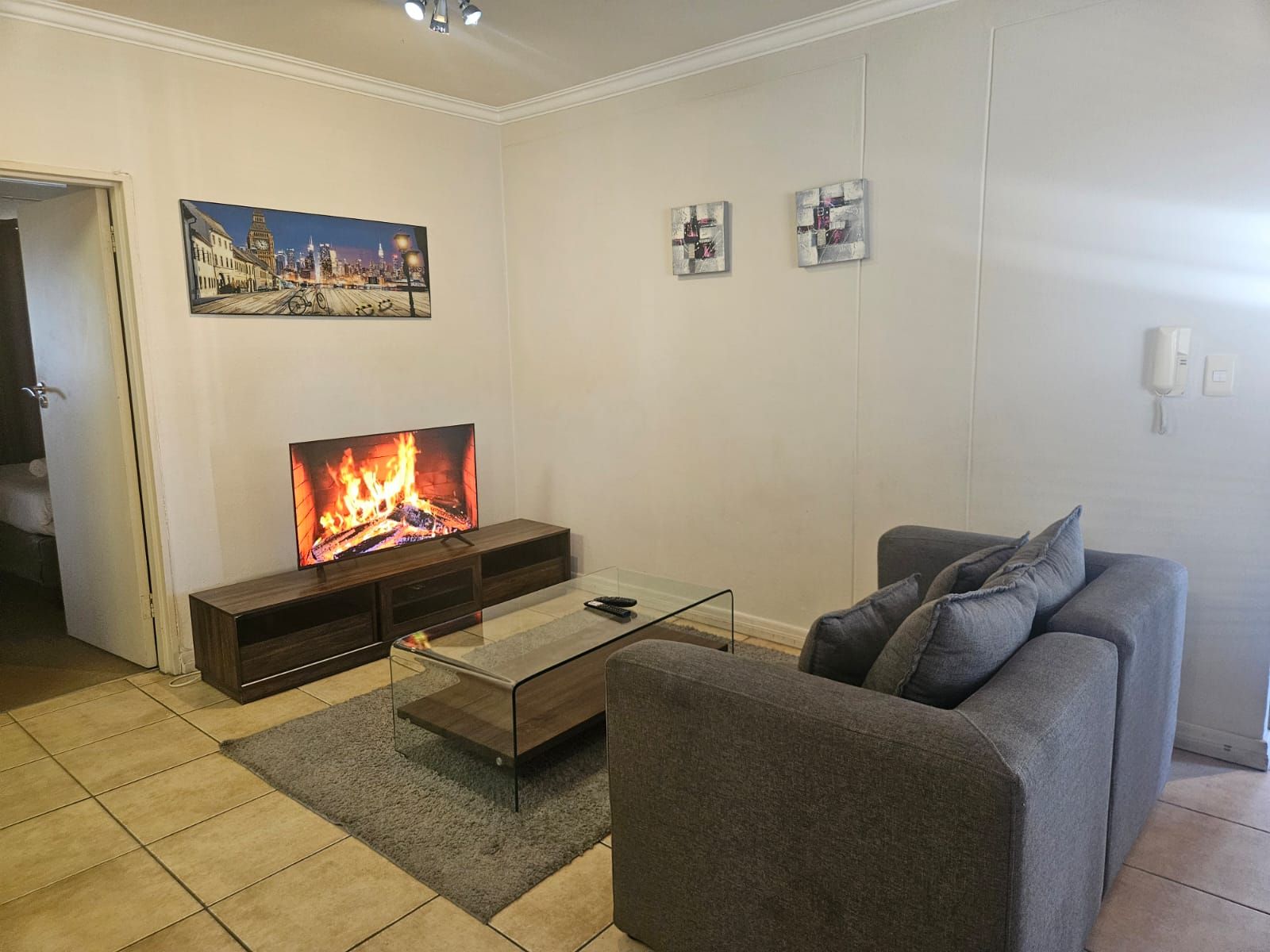 Un salón con una televisión mostrando un fuego, un sofá gris y una mesa de centro de cristal.