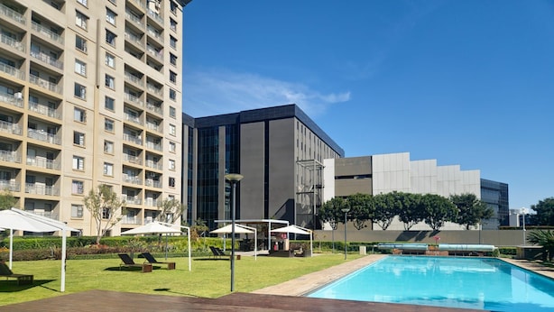 A hotel pool area with a wooden deck, a modern building, and a high-rise apartment building in the background. - Sandton Westpoint Luxury Apartments -172 (Sandton)