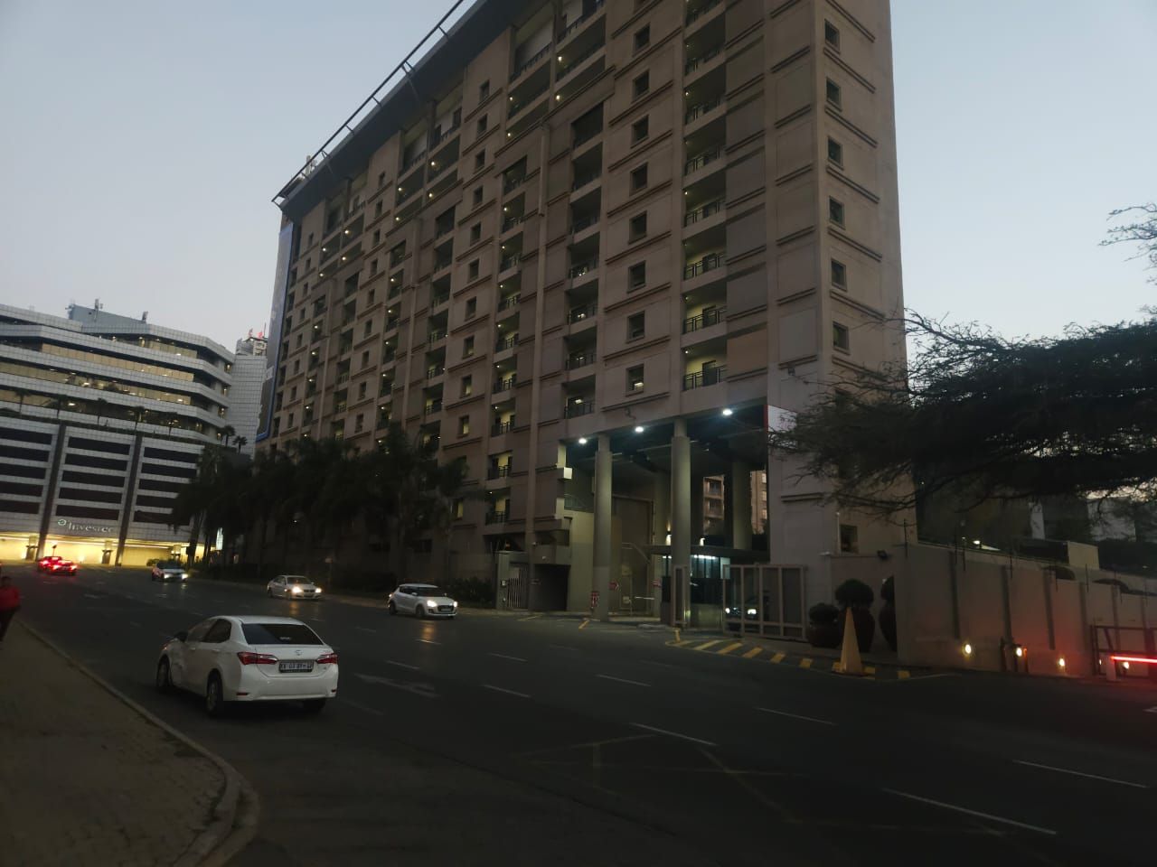 A multi-story hotel with a modern facade, a gated entrance, and a parking area with cars.