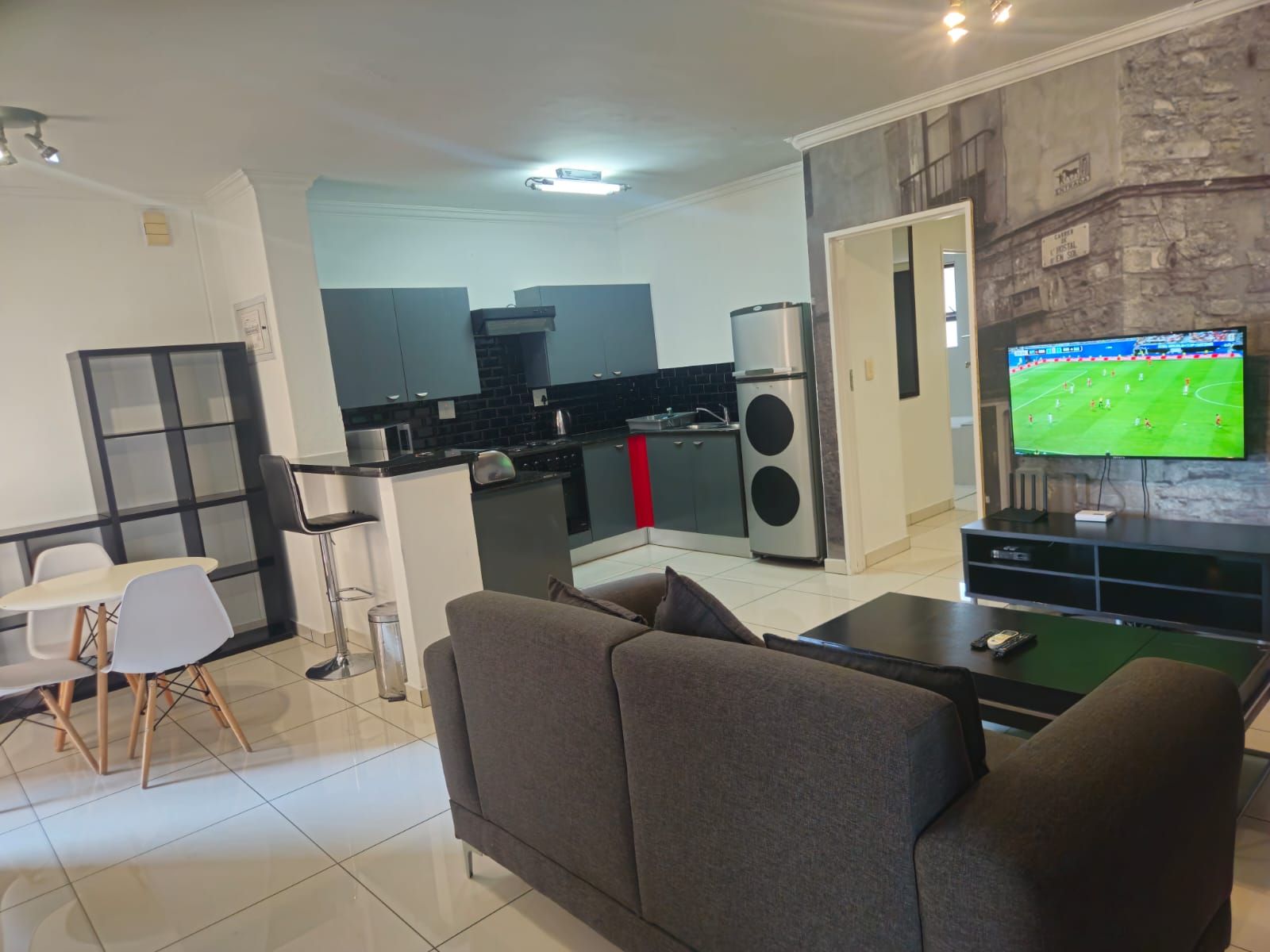 Una sala moderna con un sofá, un comedor con sillas, una cocina equipada y un televisor mostrando un partido deportivo.