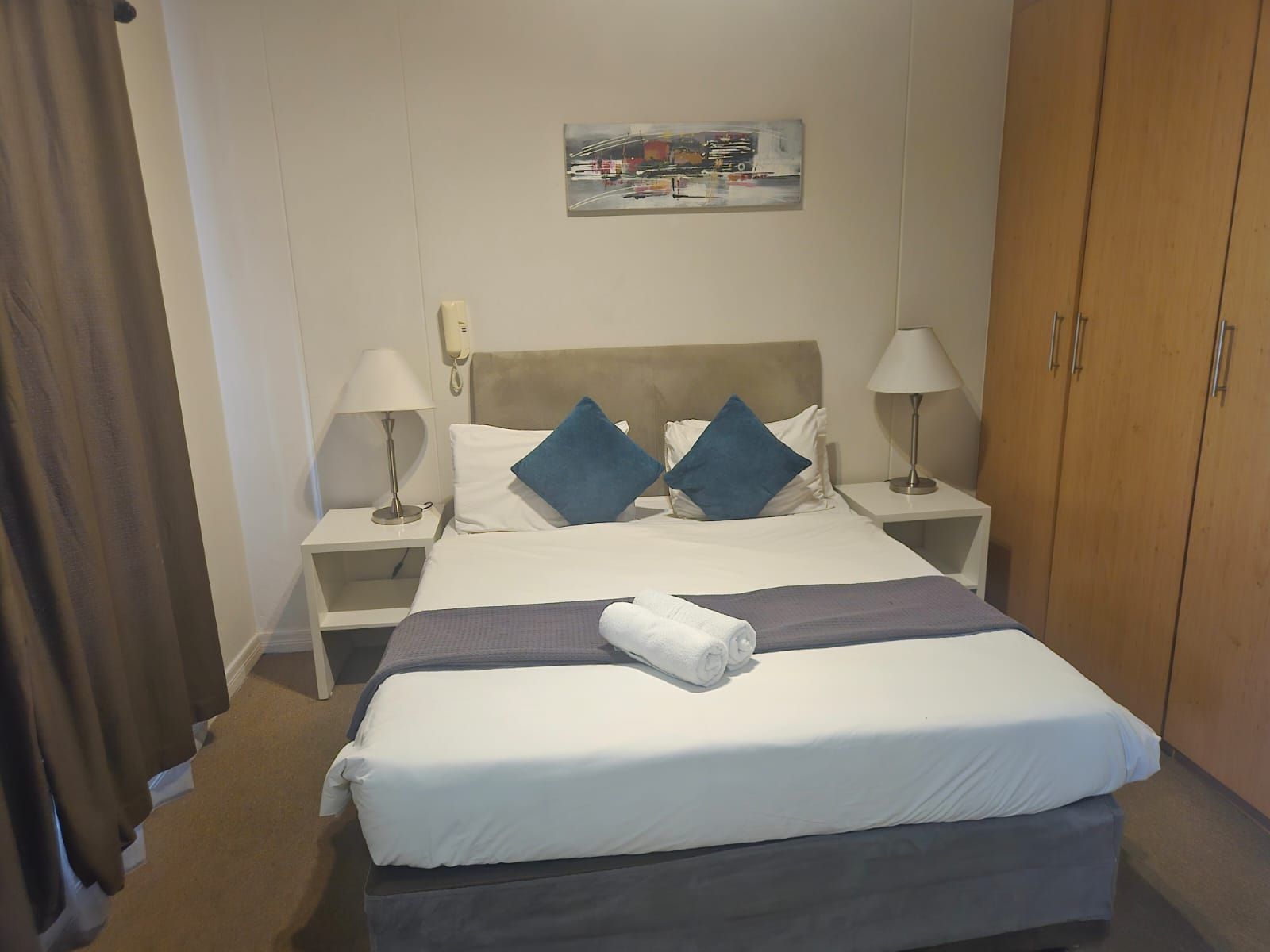 Una habitación de hotel con una cama, dos mesitas de noche, un armario y un cuadro en la pared.