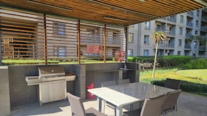 An outdoor patio with a grill, seating area, and a view of a residential building. - Sandton Westpoint Luxury Apartments -172 (Sandton)